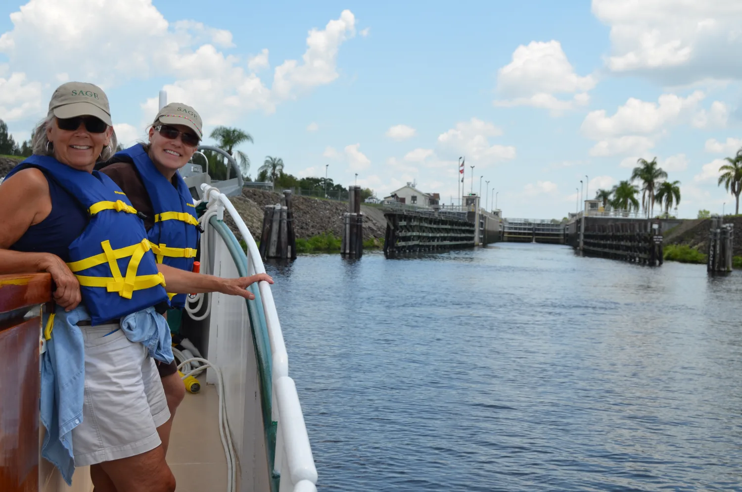 Two women in Sage caps and life jackets smiling on deck with a river lock ahead